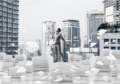 Man in casual wear keeping hand with book up while standing among flying paper planes with cityscape on background. Mixed media.