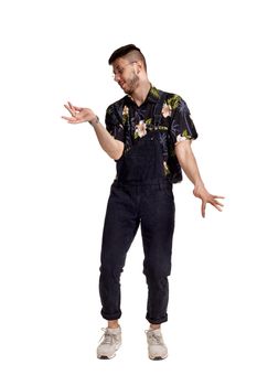 Full-length portrait of a cool person in glasses, black jumpsuit, colorful t-shirt and gray sneakers fooling around in studio. Indoor photo of a man dancing isolated on white background and looking away. Music and imagination.