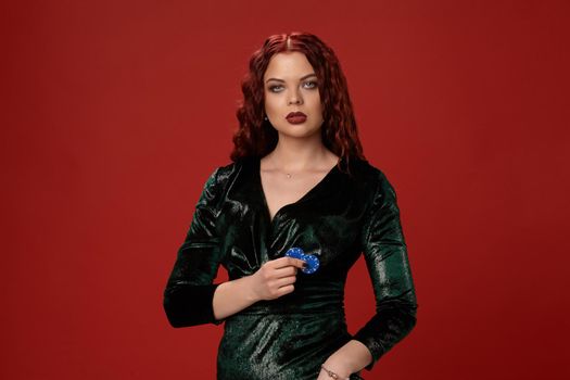 Beautiful young redheaded woman holding a blue gambling chips in her hands is posing on a red background.