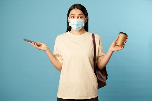 Waist up of confused young woman holding coffee and smartphone in hands while posing in studio, isolated on blue background. Copy space. Quarantine, epidemic concept