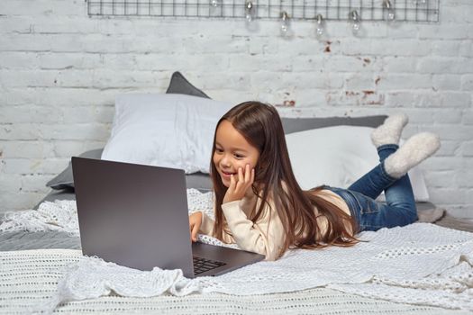 Cute little girl girl feeling amusing while watching cartoons on a laptop sitting on bed. White sweater and jeans.