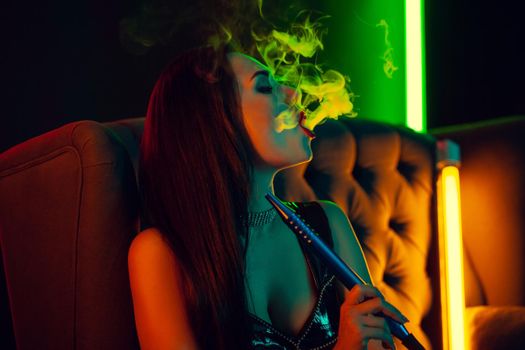 Close-up shot of a pretty brunette girl with a perfect make-up, in a sparkling necklace, leather dress and patent boots smoking a hookah exhaling a smoke sitting on an arm-chair at a luxury night club. Concept of pleasant spending time. Dark background with colorful neon backlights.