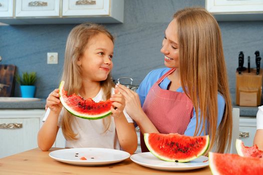 Mother and daughter eating watermelon at home in kitchen