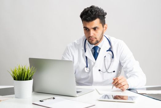 Young arab internist working at computer. Physician in white coat with stethoscope sitting at desk with laptop. Professional medical consultation and treatment in modern clinic. Medical healthcare