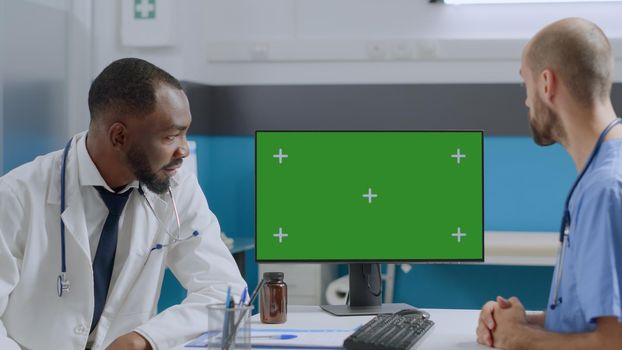 Multi-ethnic team looking at mock up green screen chroma key computer with isolated display discussing healthcare treatment in hospital office. African american doctor explaining sickness expertise