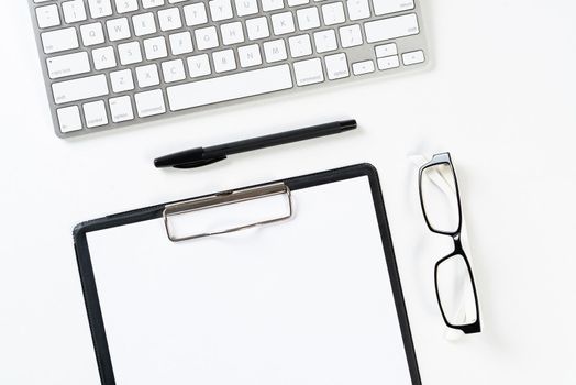 Top view of well organized workplace with office accessories. Clipboard with blank paper page, glasses and wireless keyboard on white surface. Education, creativity and working concept with copy space