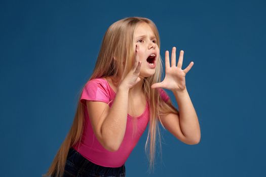 Portrait of adorable blonde little girl on blue background. in pink shirt shouting, trying to call someone