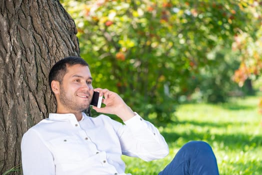 Young man talking on smartphone under tree in park on sunny day. Handsome businessman relaxing outdoors. Smiling man sitting on green grass leaning against tree. Mobile communication concept.