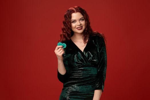 Beautiful redheaded woman holding a blue gambling chips in her hand and smiling, on a red background.