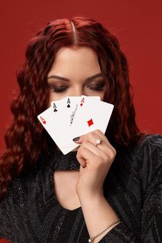 Beautiful redheaded woman in a black knitted dress have covered her face with some playing cards and closed her eyes. Casino. Four aces. Studio shot on a red background.