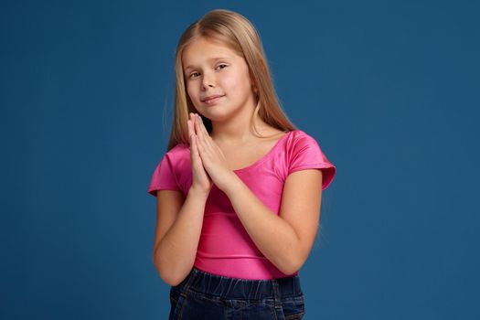 Portrait of adorable blonde little girl on blue background. in pink shirt, pretending cute innocent person and smiling
