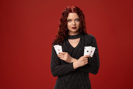 Beautiful redheaded woman in a black knitted dress is holding some playing cards and looks serious. Casino. Four aces. Studio shot on a red background.