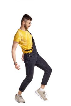 Full-length portrait of a sporty male in glasses, black jumpsuit, yellow t-shirt and gray sneakers fooling around in studio. Indoor photo of a man making dancing elements sideways isolated on white background. Music and imagination concept.