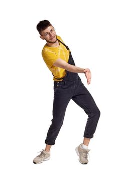 Full-length portrait of an elegant guy in glasses, black jumpsuit, yellow t-shirt and gray sneakers fooling around in studio. Indoor photo of a man making dancing elements isolated on white background. Music and imagination. Copy space.