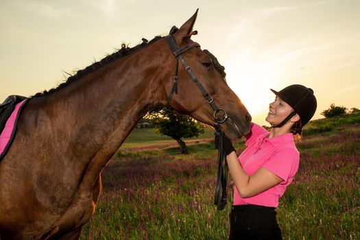Beautiful smiling girl jockey stand next to her brown horse wearing special uniform on a sky and green field background on a sunset. Equitation sport competition and activity.