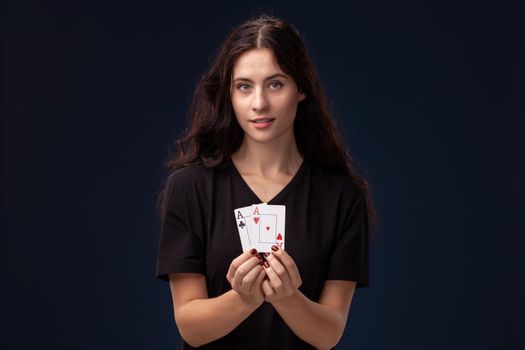 Curly hair brunette wearing a blackish t-shirt is posing with playing cards in her hands and looking at the camera. Poker concept on a black background. Casino.