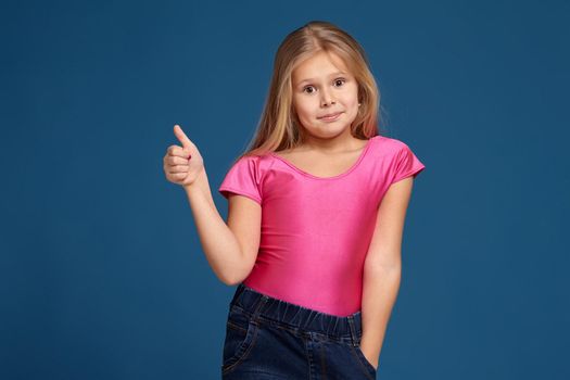 Close-up emotional portrait of caucasian little girl in pink shirt on blue background, making thumbs up gesture, looks happy