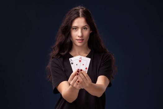 Curly hair brunette wearing a blackish t-shirt is showing playing cards in her hands and looking at the camera. Poker concept on a black background. Casino.