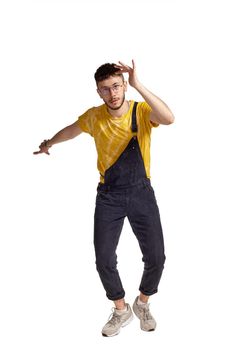 Full-length portrait of a fasionable guy in glasses, black jumpsuit, yellow t-shirt and gray sneakers fooling around in studio. Indoor photo of a man making dancing elements isolated on white background. Music and imagination concept.
