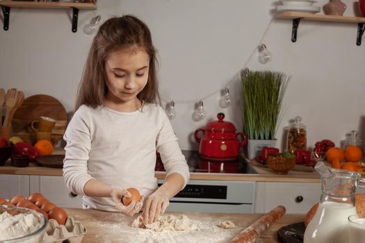 Charming little brunette girl dressed in a white blouse is kneading the dough for baking a bread and holding an egg in her hand at a kitchen against a white wall with shelves and bulbs on it. Happy childhood concept.
