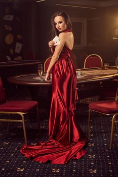 Young brunette girl in a long burgundy atlas dress, with two aces in her hand is posing sideways near a poker table in vip casino. Passion, cards, chips, alcohol drinks, win, gambling - it is a best entertainment.