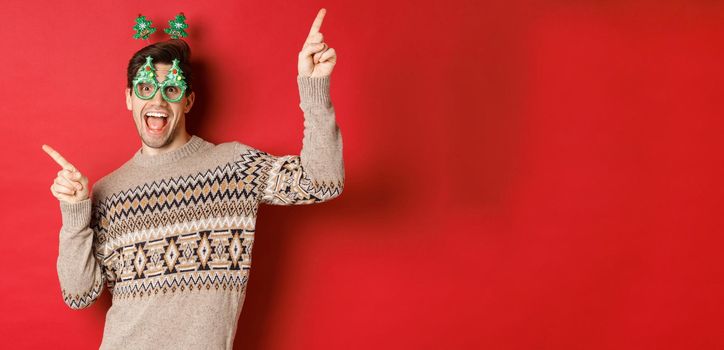 Portrait of joyful handsome guy in party glasses and christmas sweater, dancing and pointing fingers sideways, enjoying new year celebration, standing over red background.