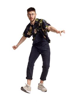 Full-length portrait of a graceful male in glasses, black jumpsuit, colorful t-shirt and gray sneakers fooling around in studio. Indoor photo of a man dancing isolated on white background and looking at the camera. Music and imagination.