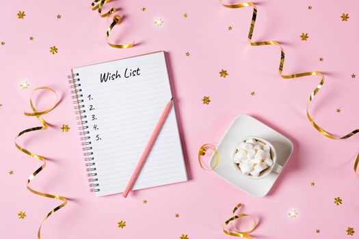New year, Christmas or holiday wish list concept. Notepad, golden tinsel, and a cup of hot chocolate with marshmallows isolated on pink background. Banner with copy space