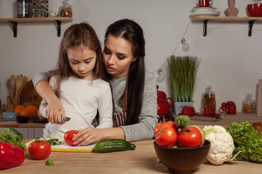Happy loving family are cooking together. Pretty mother and her little princess are making a vegetable salad and having fun at the kitchen, against a white wall with shelves and bulbs on it. Homemade food and little helper.