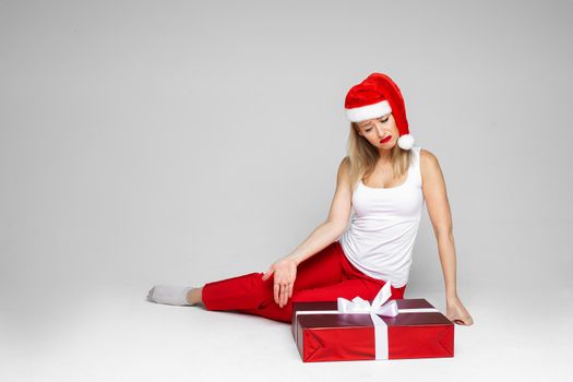 Disappointed blonde lady wearing Santa hat while sitting near big red Christmas present. Copy space