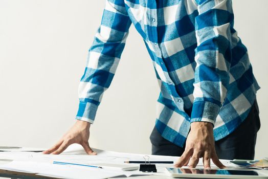 Architect standing near desk with paper blueprints. Architecture studio concept with design project. Close up man hand lying on construction drawing. Professional building, engineering and renovation