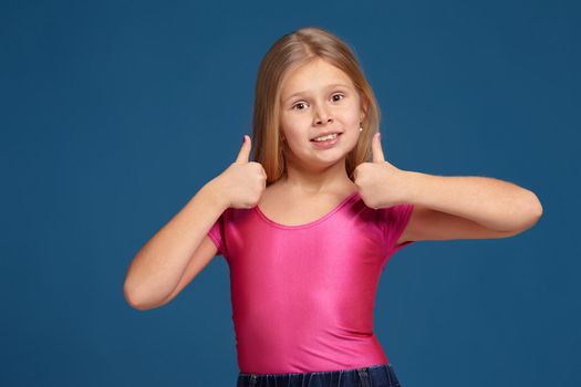 Close-up emotional portrait of caucasian little girl in pink shirt on blue background, making thumbs up gesture, looks happy