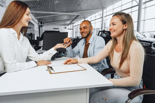 Cheerful couple taking keys of their new car in car dealership from saleswoman