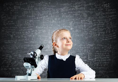 Little girl scientist with microscope on chalkboard background. Research and discovery concept with copy space. Elementary science class in modern school. Schoolgirl in schoolwear sitting at desk.