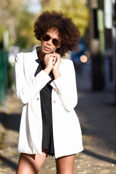Young black woman with afro hairstyle standing in urban background with aviator sunglasses. Mixed girl wearing white jacket and black dress posing near a brick wall