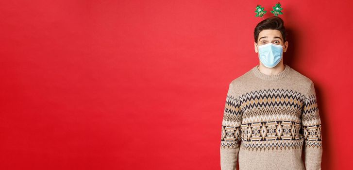 Concept of christmas, covid-19 and social distancing. Cheerful young man in medical mask and new year clothing, celebrating holidays during pandemic, standing over red background.