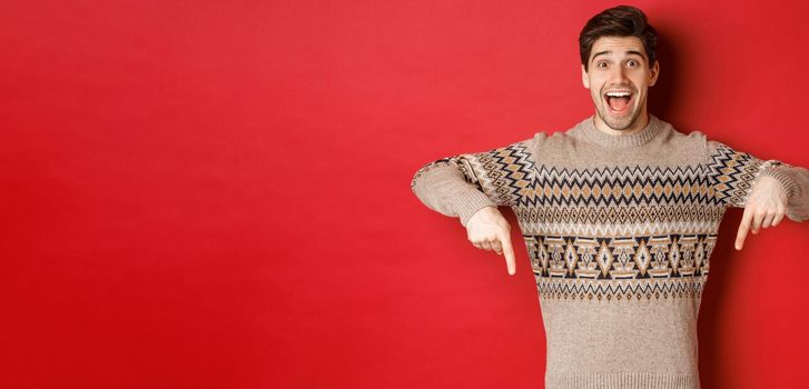 Concept of christmas celebration, winter holidays and lifestyle. Excited caucasian male model in xmas sweater, pointing fingers down and making announcement, showing advertisement, red background.