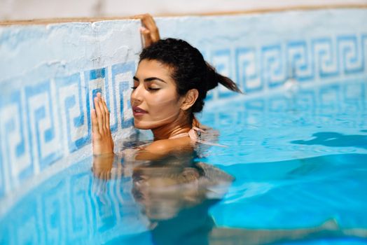 Beautiful Arab woman relaxing in swimming pool with eyes closed. Girl with healthy tanned skin and wet hair enjoying Summer Sun.