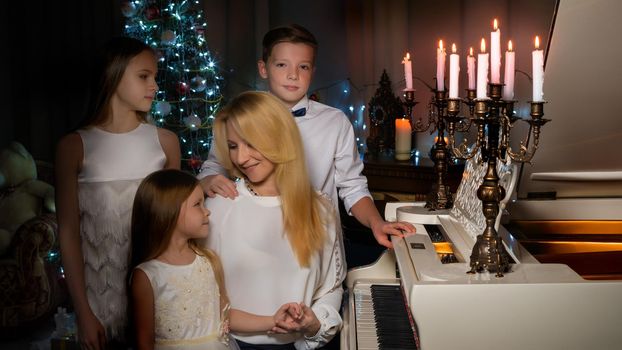 A happy mother with young children on Christmas Eve in a dark room with candles and a fireplace, near a large white grand piano. The concept of a family holiday.