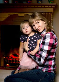 A young mother with a little girl by the fireplace on Christmas Eve. The concept of family happiness, a holiday.