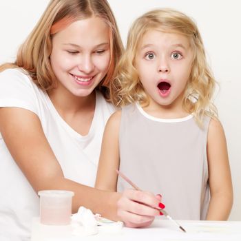 The older sister paints with the younger sister paints at the table on a piece of paper.