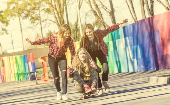 Girls pushing friend on the skateboard at sunny day. Cute young girls having fun outdoors