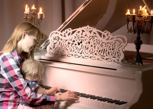 A young mother with a small child on Christmas night with candles near a large white piano. The concept of holidays and family happiness.