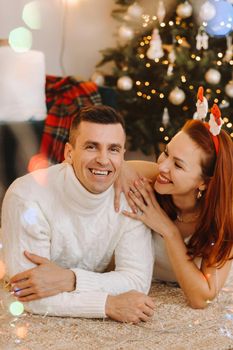 a happy married couple is lying on the floor near the Christmas tree at home.