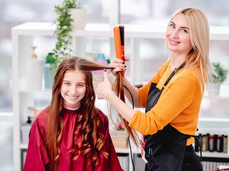 Hairdresser making beautiful curls with a hair iron for smiling beautiful young model girl