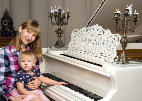 A young mother with a small child on Christmas night with candles near a large white piano. The concept of holidays and family happiness.