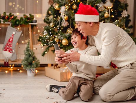 Happy father and little son blowing up festive petard while sitting under decorated christmas tree at home