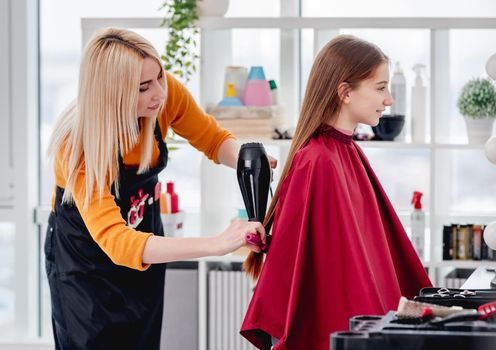 Beautiful blond hairdresser straightens hair of young girl model using hairdryer