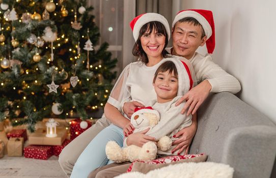 Happy family in santa hats sitting on sofa and hugging next to illuminated christmas tree at home
