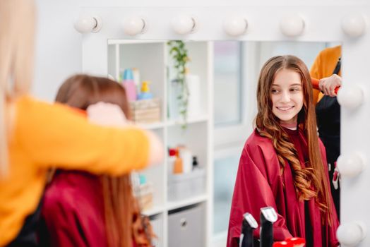Reflection in mirror of smiling attractive young model with curls during hairstyle process in beauty salon